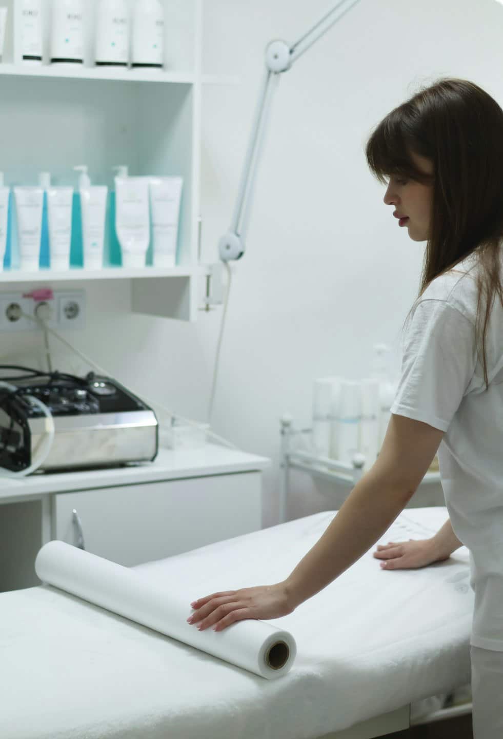 An aesthetician unrolls white paper onto a spa treatment bed.
